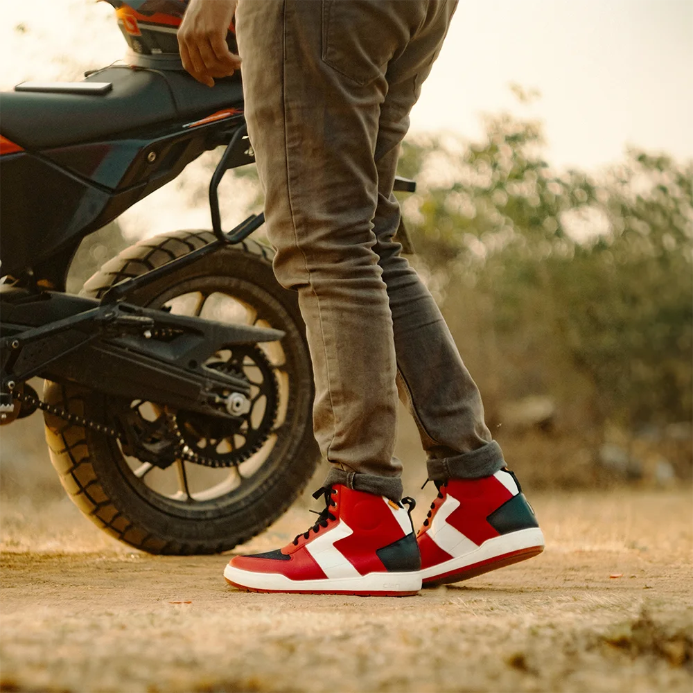 Person wearing white red D30 Stealth Shoes standing next to a motorcycle on a dirt road.